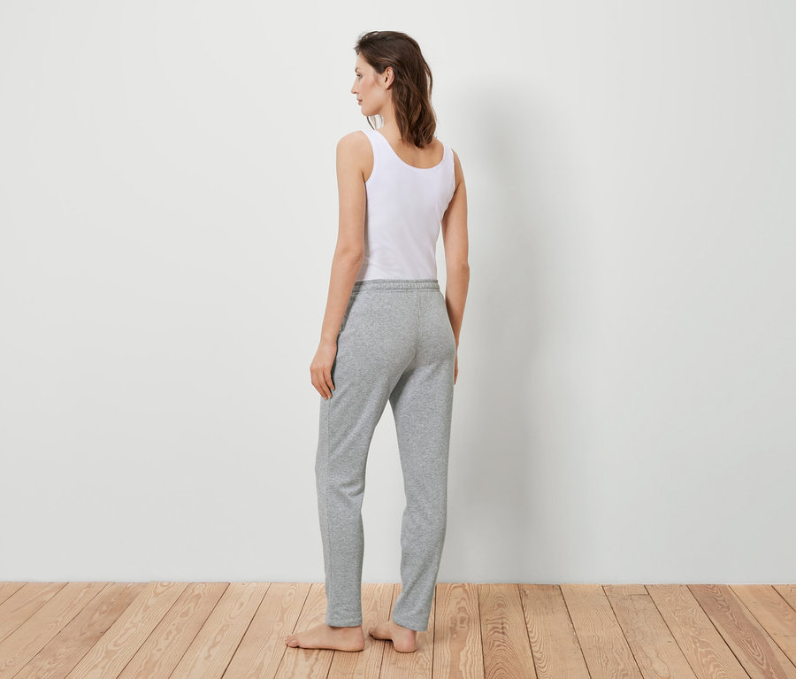 Frau steht mit dem Rücken zur Kamera, trägt ein weißes Tanktop und graue Jogginghose.