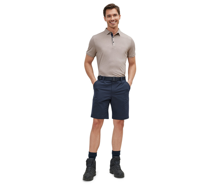 Mann steht mit den Händen in den Taschen, trägt ein beiges Poloshirt und eine blaue 2-in-1-Funktionshose.