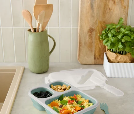 Eine blaue, unterteilte Lunchbox mit Brombeeren, Nüssen und Couscous mit Gemüse steht auf der Theke.