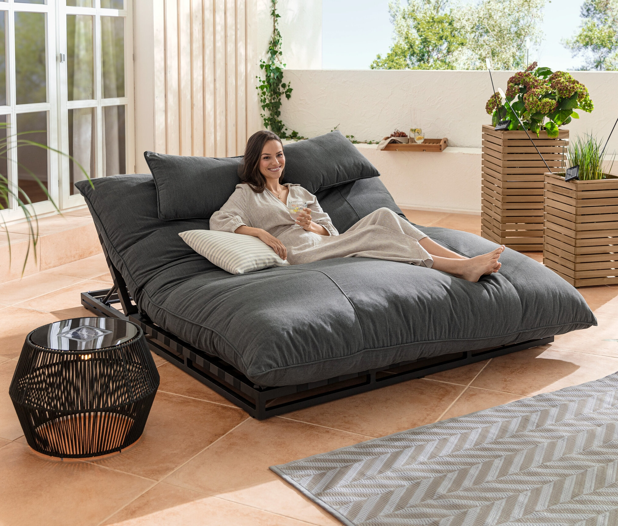 Frau liegt auf Premium-Doppelliege mit verstellbarem Kopfteil neben Solar-Beistelltisch mit Textilband und Pflanzsäulen-Set »Tinus«.
