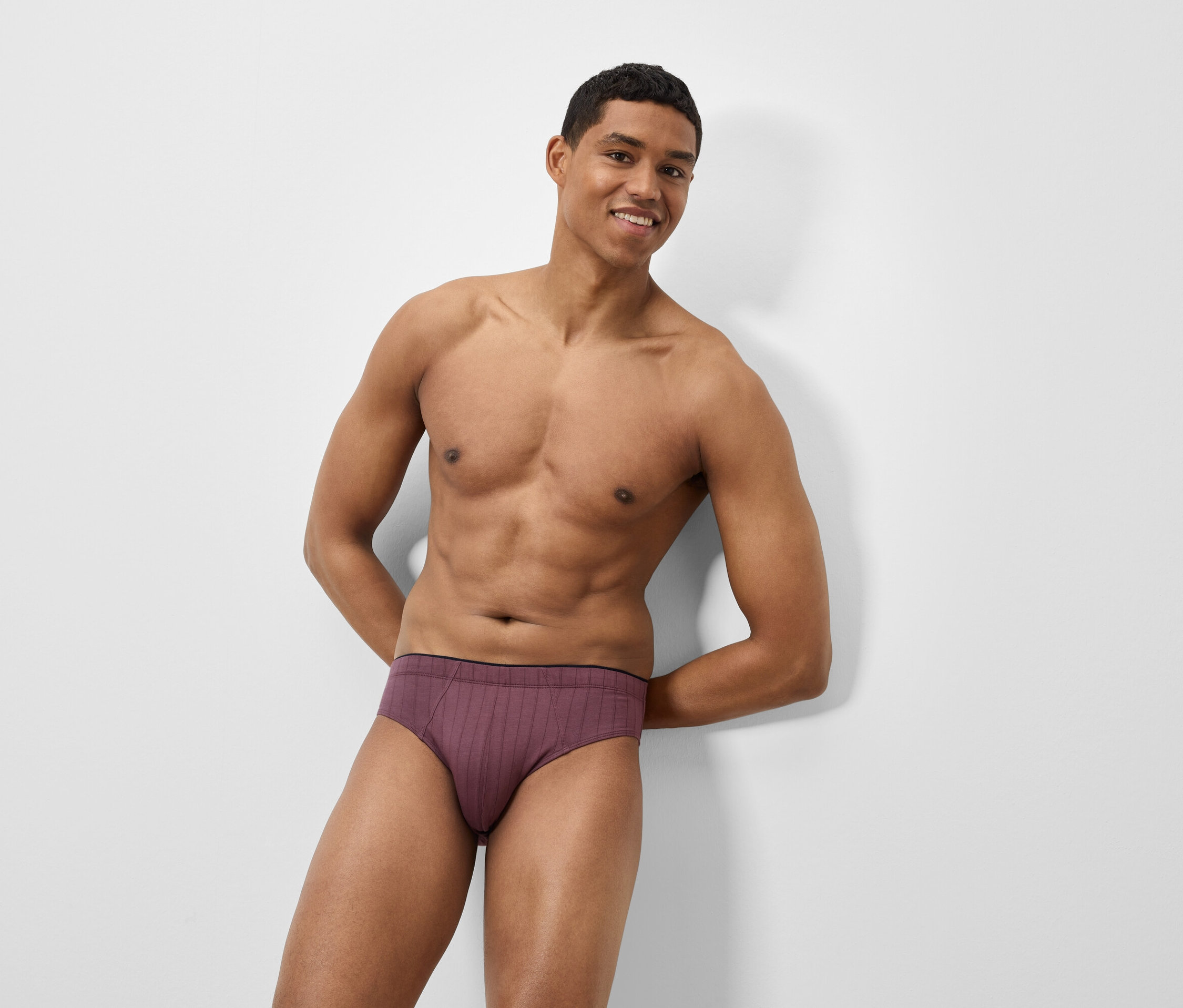 Ein braunhäutiger Mann lehnt an einer weißen Wand und trägt weinrote Slips.