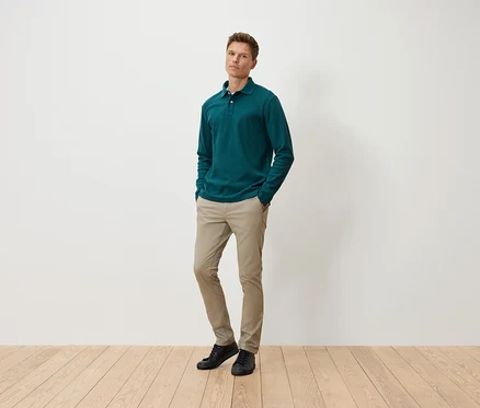 Ein Mann steht mit den Händen in den Hosentaschen und trägt ein grünes Langarm-Poloshirt, beige Hosen und schwarze Schuhe.