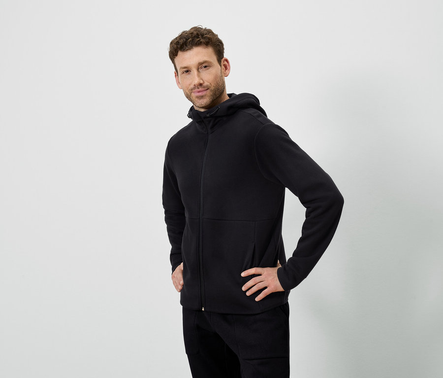 Ein Mann posiert vor einem weißen Hintergrund. Er trägt einen schwarzen Kapuzenpullover und eine schwarze Hose.