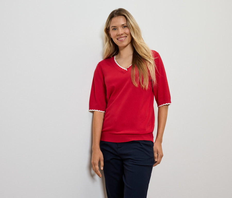 Eine Frau mit blonden Haaren posiert in einem roten Pullover und dunkelblauen Hosen.