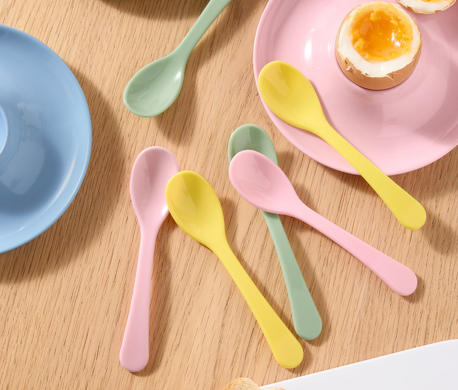 Draufsicht auf einen Tisch mit einem gekochten Ei in einem Eierbecher, einem halbierten gekochten Ei auf einem Teller, farbigen Löffeln und Toast.