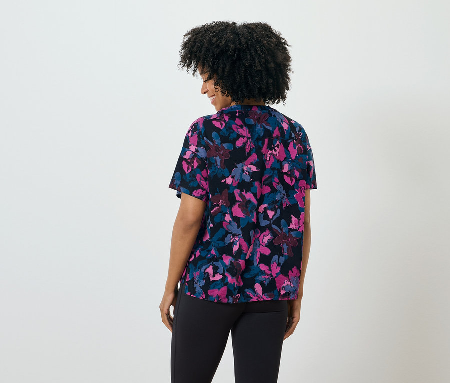 Frau mit dunklen Haaren von hinten trägt eine schwarze Bluse mit Blumenmuster und eine schwarze Hose.