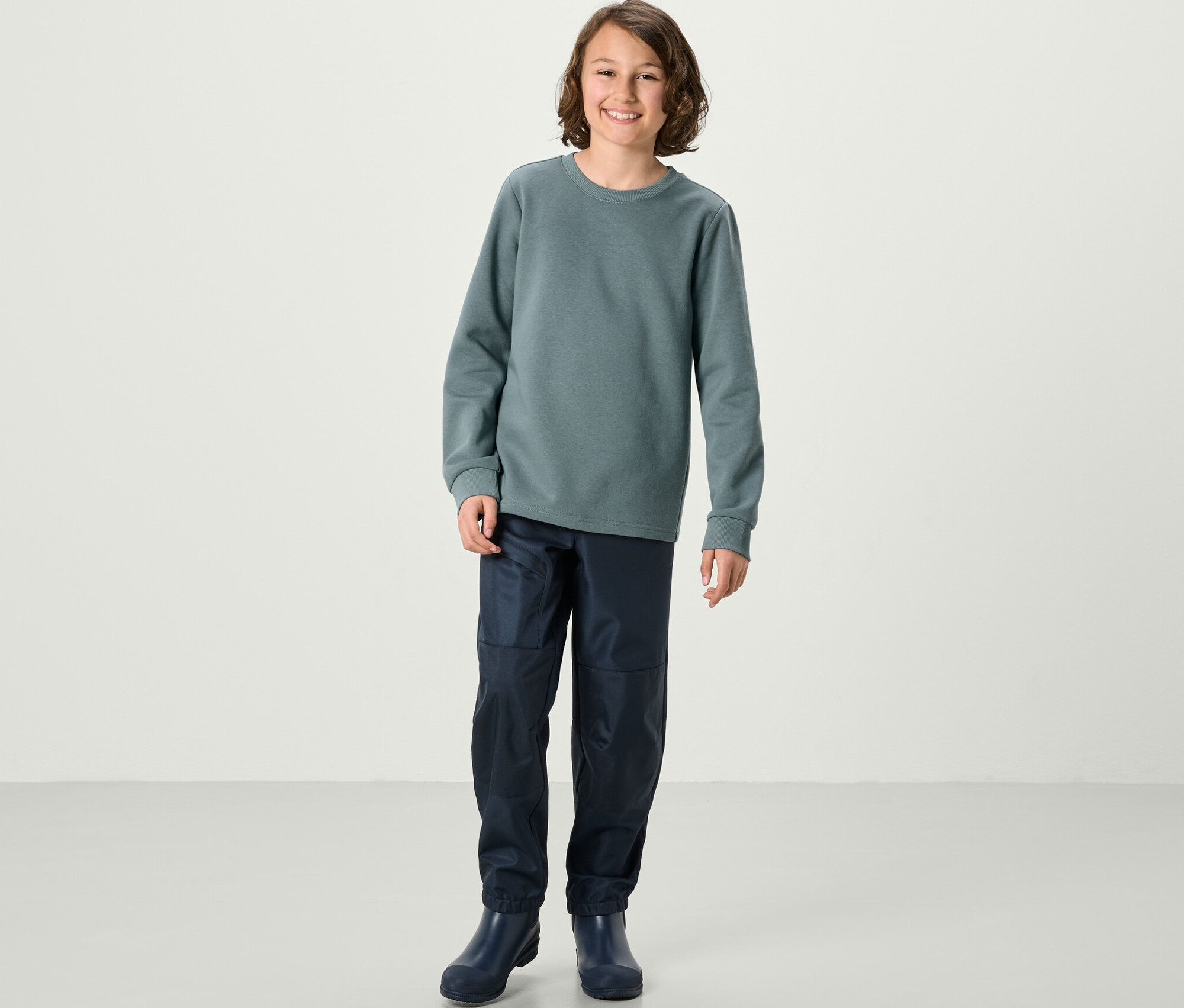 Ein Junge steht und trägt ein blaues Sweatshirt und Hosen. 2 Kinder-Sweatshirts mit angerauter Innenseite, blau und salbei.