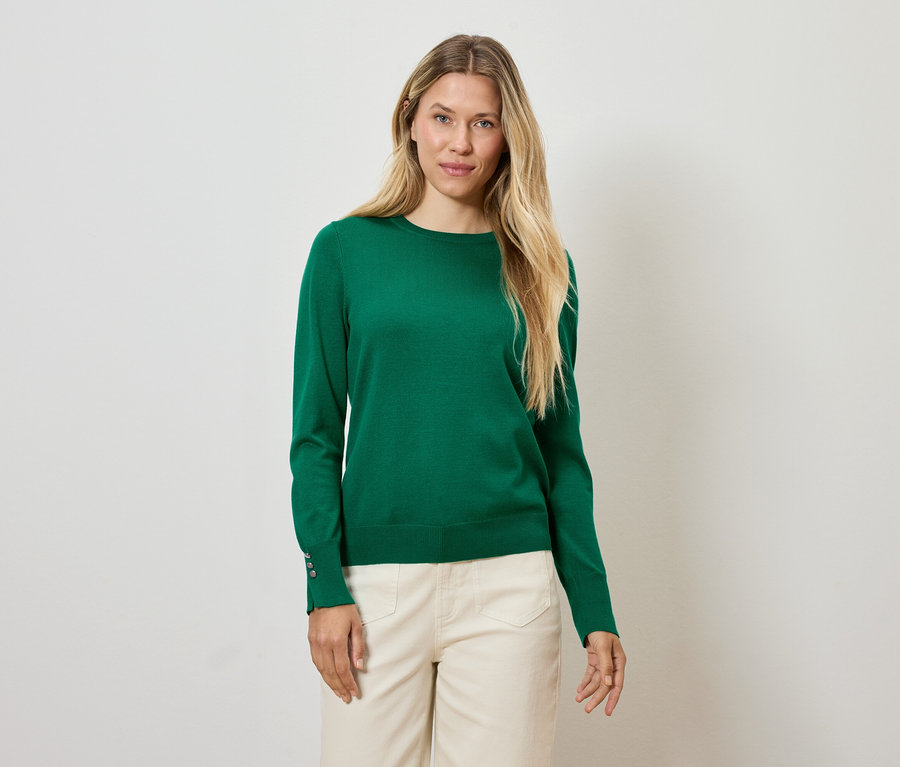 Blonde Frau in grünem Pullover und cremefarbener Hose steht vor einem weißen Hintergrund.