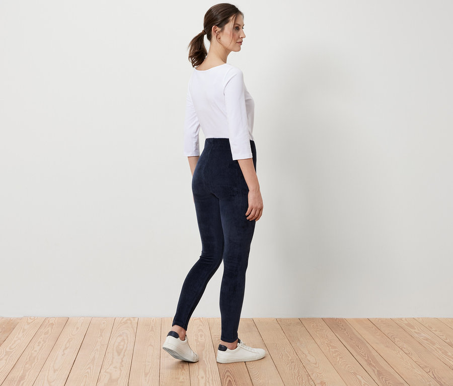 Frau in weißem Oberteil und blauen Cord-Leggings steht auf Holzboden.