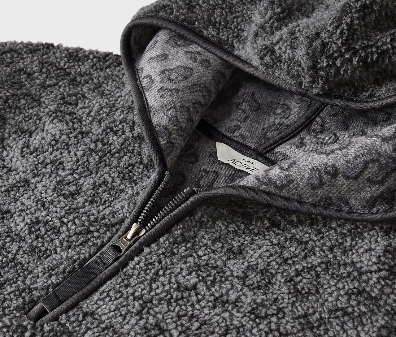 Detailansicht eines grauen, zotteligen Kapuzenpullovers mit Reißverschluss und grauem Tiermuster auf der Innenseite der Kapuze.