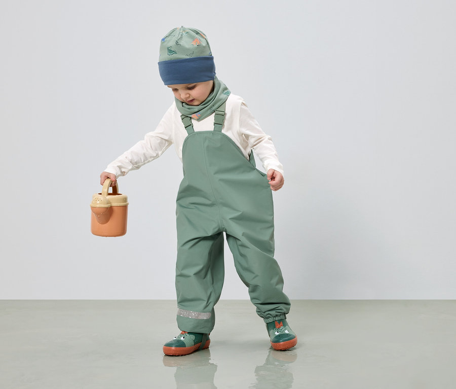 Ein Kind in grüner Latzhose, Mütze und Halstuch mit Dinosaurier-Muster hält eine Gießkanne in der Hand.