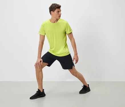 Mann in Sportbekleidung dehnt sich. Er trägt ein neon-grünes T-Shirt, schwarze Shorts und schwarze Turnschuhe.