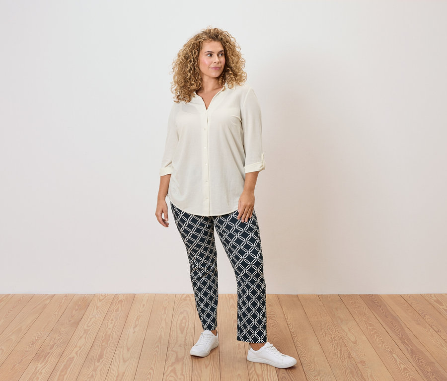 Frau mit lockigem Haar trägt eine cremefarbene Bluse, gemusterte Hosen und weiße Turnschuhe.