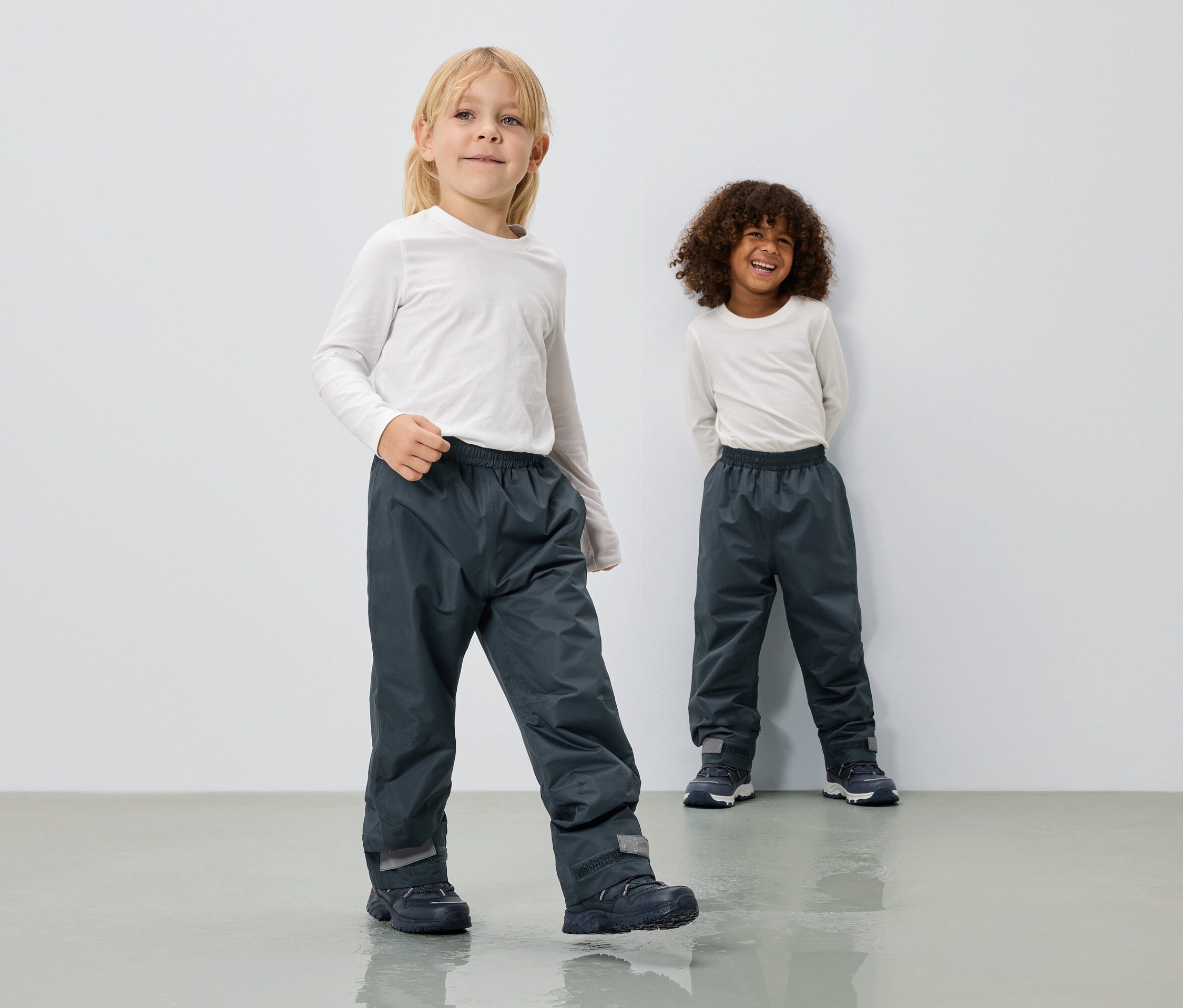 Zwei Kinder tragen lange, graue Regenhosen und langärmlige, weiße Oberteile vor einer weißen Wand.