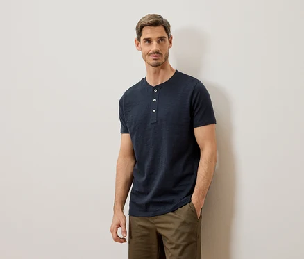 Mann mit blauem Henley-Shirt und khakifarbenen Hosen, der an einer weißen Wand posiert.