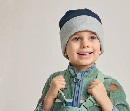 Lächelnder Junge mit blau-grauer Mütze und grünem Dino-Overall hält Hosenträger.