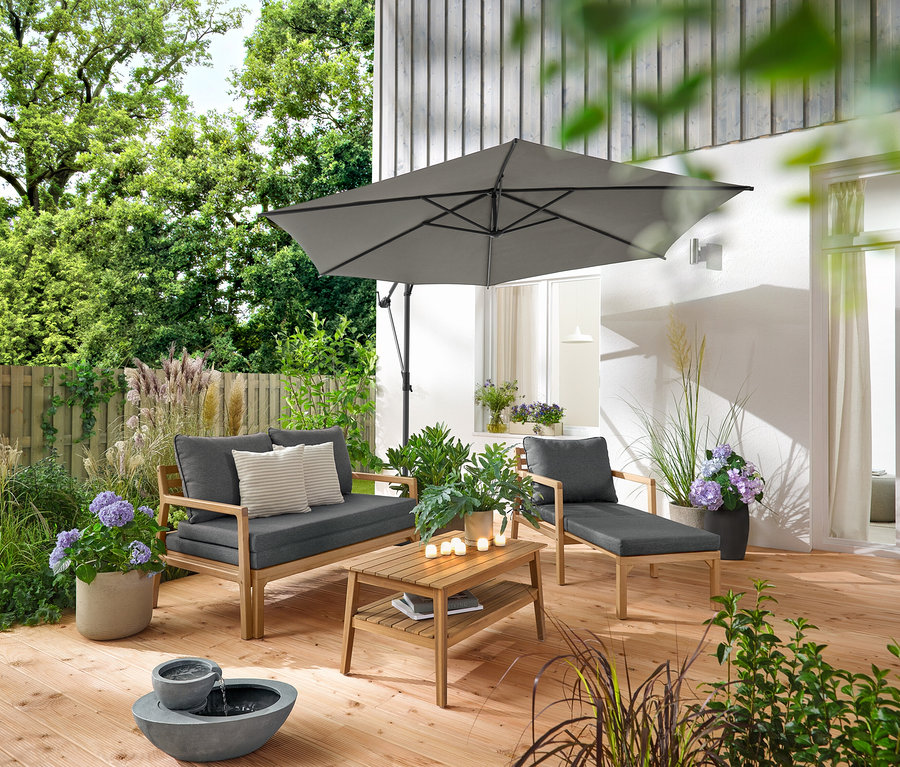 Auf einer Holzterrasse stehen ein ausziehbarer Gartensessel, ein ausziehbares Gartensofa, ein Garten-Couchtisch, ca. 80 x 50 cm, und ein Solar-Gartenbrunnen. Ein anthrazitfarbener Leco Ampelschirm spendet Schatten. 2 Outdoor-Kissen mit Leinen-Haptik und 6 LED-Outdoor-Akku-Teelichter dekorieren den Bereich.