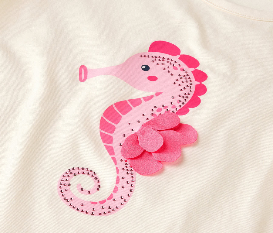 Nahaufnahme eines hellen Stoffes mit einem rosa Seepferdchen-Design, das mit Strasssteinen und einer Stoffblume verziert ist.
