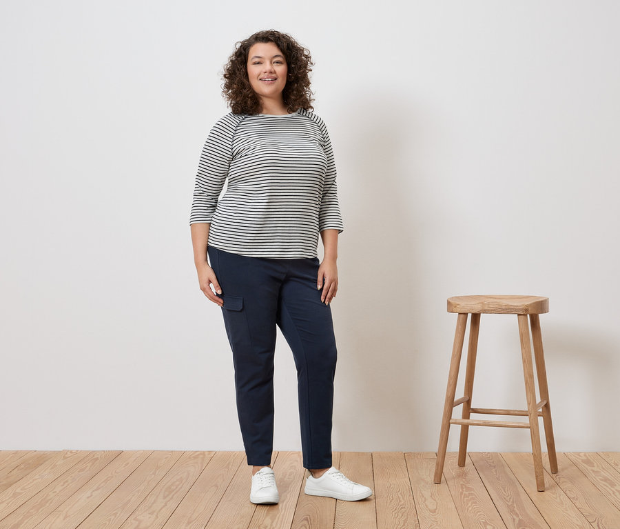 Frau mit lockigem Haar steht in einem gestreiften Oberteil, blauen Hosen und weißen Turnschuhen neben einem Hocker.