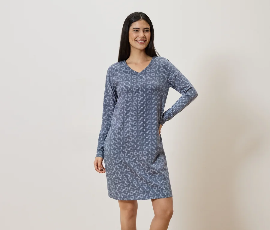 Lächelnde Frau mit dunklen Haaren trägt ein blaues, gemustertes, langärmeliges Kleid.