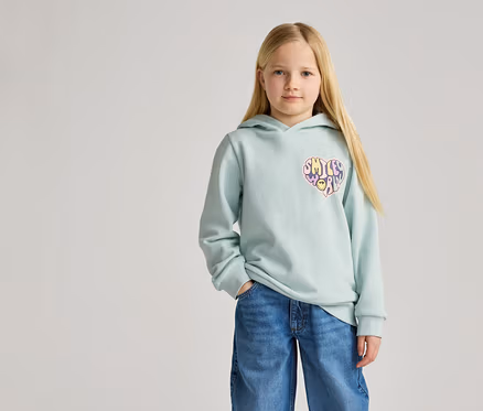 Mädchen mit blonden Haaren trägt hellblauen Kapuzenpullover mit Herz-Smiley-Aufdruck und Jeans.