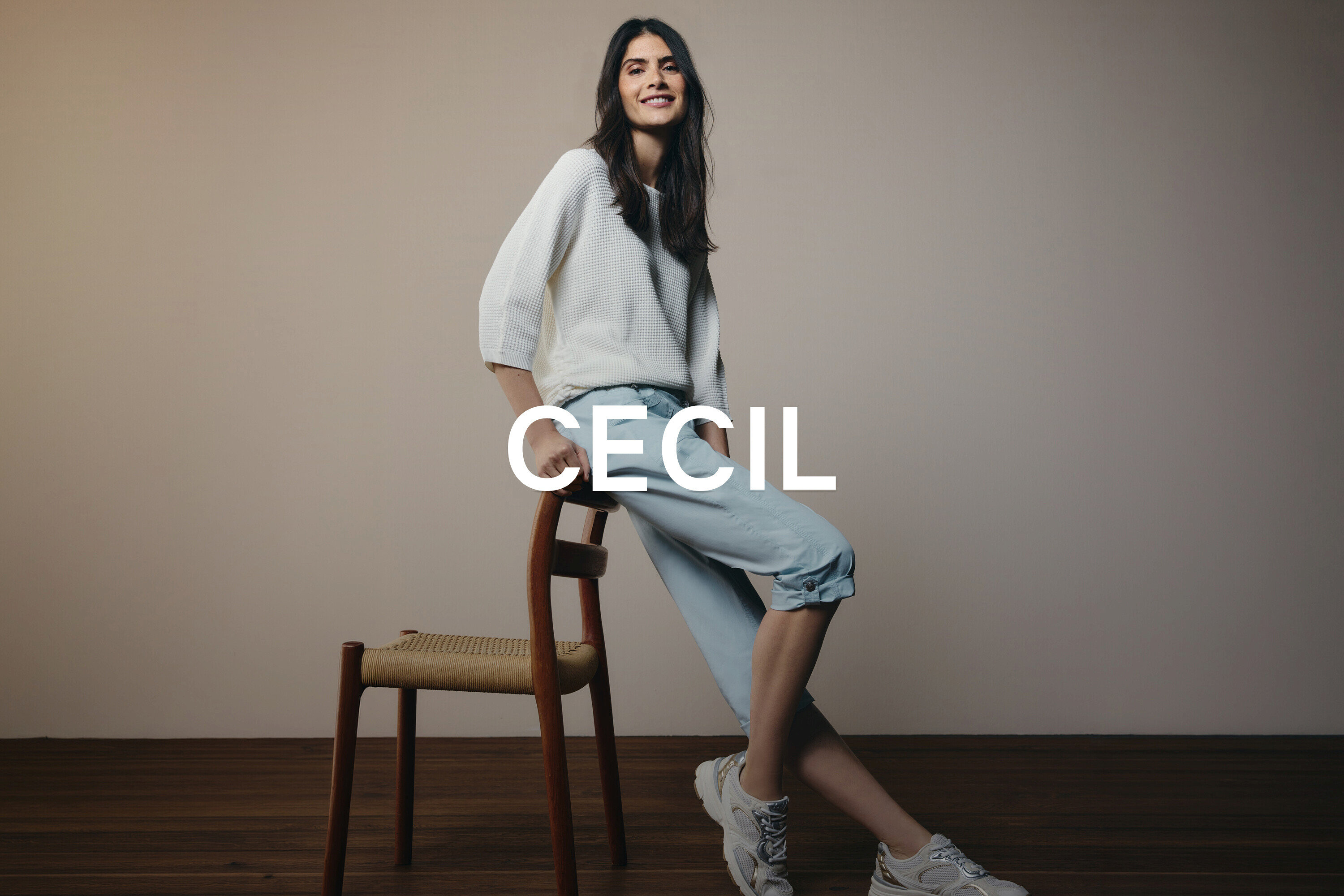 Frau sitzt auf einem Holzstuhl, trägt einen weißen Pullover, hellblaue Jeans und weiße Turnschuhe.