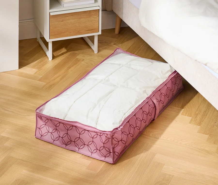 Auf einem Holzboden liegt eine rosafarbene Aufbewahrungstasche unter einem Bett neben einem Nachttisch.
