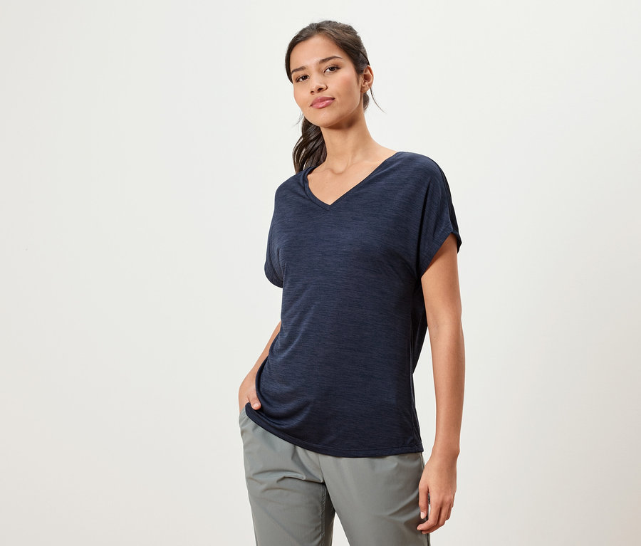 Frau mit dunklem T-Shirt und grauer Hose steht vor einem weißen Hintergrund.