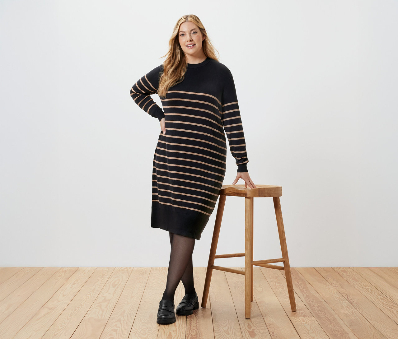 Frau posiert in einem gestreiften Strickkleid neben einem Hocker.