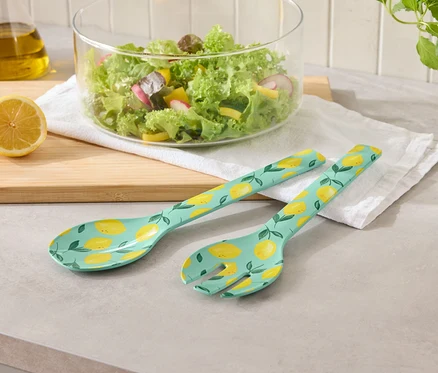 Salatbesteck mit Zitronenmuster neben Schüssel Salat und Zitronen auf einem Tisch.