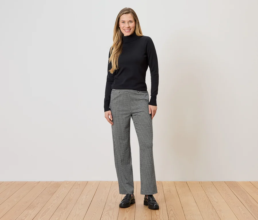 Eine Frau steht vor einer Wand und trägt einen schwarzen Rollkragenpullover und eine schwarz-weiß karierte Hose.