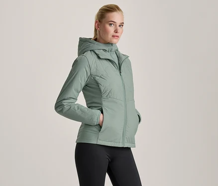 Frau in einer hellgrünen Kapuzenjacke und schwarzen Leggings, die mit den Händen in den Jackentaschen steht.