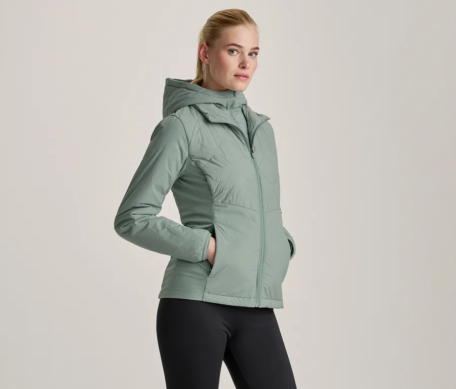 Frau in einer hellgrünen Kapuzenjacke und schwarzen Leggings, die mit den Händen in den Jackentaschen steht.