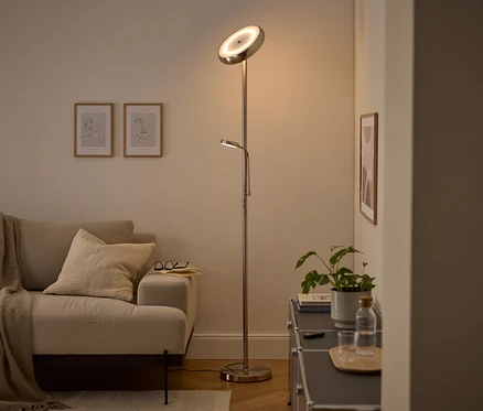 Eine moderne silberne Stehlampe mit nach oben gerichtetem Hauptlicht und verstellbarer Leselampe beleuchtet ein gemütliches Wohnzimmer. Ein beiges Sofa mit Decke und Kissen, zwei gerahmte Line-Art-Bilder und ein dunkler Schrank mit Pflanze und Wasserflasche sind sichtbar.