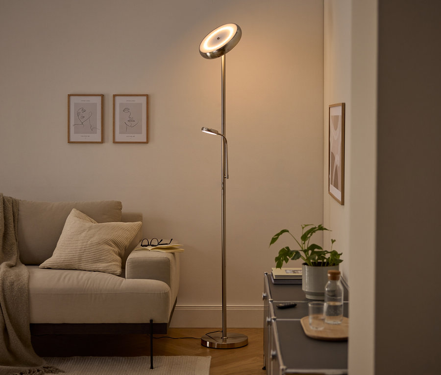 Eine moderne silberne Stehlampe mit nach oben gerichtetem Hauptlicht und verstellbarer Leselampe beleuchtet ein gemütliches Wohnzimmer. Ein beiges Sofa mit Decke und Kissen, zwei gerahmte Line-Art-Bilder und ein dunkler Schrank mit Pflanze und Wasserflasche sind sichtbar.