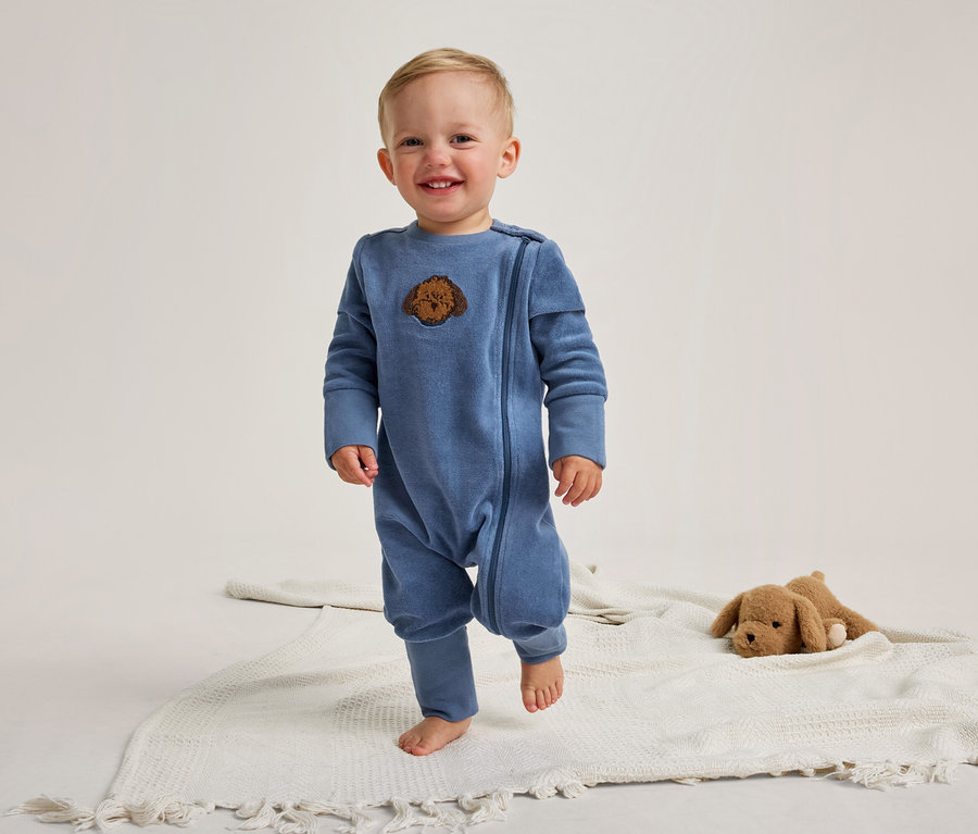 Ein lächelndes Baby steht barfuß auf einer weißen Decke. Es trägt einen blauen Strampler mit einem braunen Hundekopf-Aufnäher. Rechts liegt ein braunes Plüschtier.