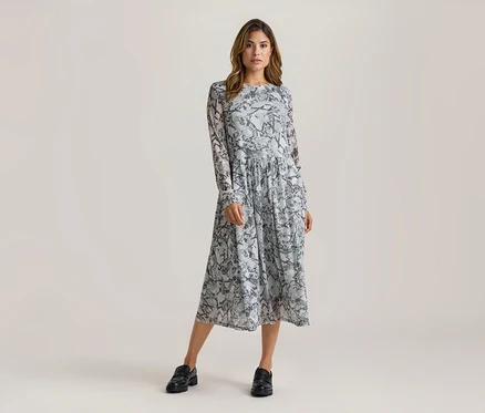 Frau steht in einem langärmeligen Midikleid mit grau-schwarzem Schlangenmuster und schwarzen Loafern.