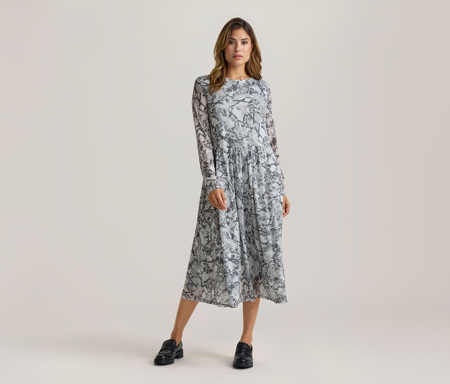 Frau steht in einem langärmeligen Midikleid mit grau-schwarzem Schlangenmuster und schwarzen Loafern.