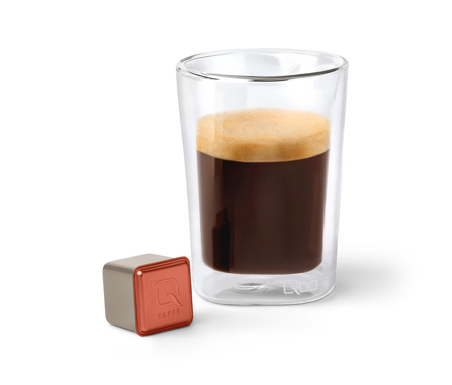 Eine Qbo Caffè TERRENO ALTO Kapsel neben einem Glas Kaffee.