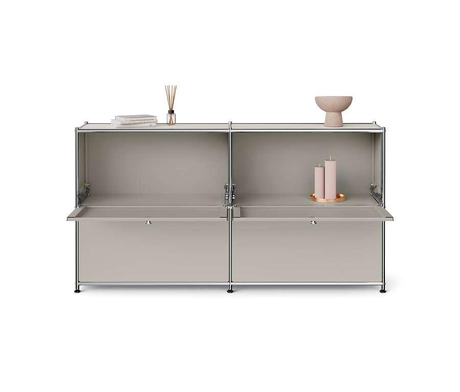 Hellgrauer modularer Schrank mit Chromrahmen, mit zwei offenen oberen Fächern und zwei offenen, herunterklappbaren unteren Türen. Auf den oberen Ablagen sind Bücher, ein Duftdiffusor, eine Holzschale und zwei rosa Kerzen auf einem goldenen Tablett zu sehen.