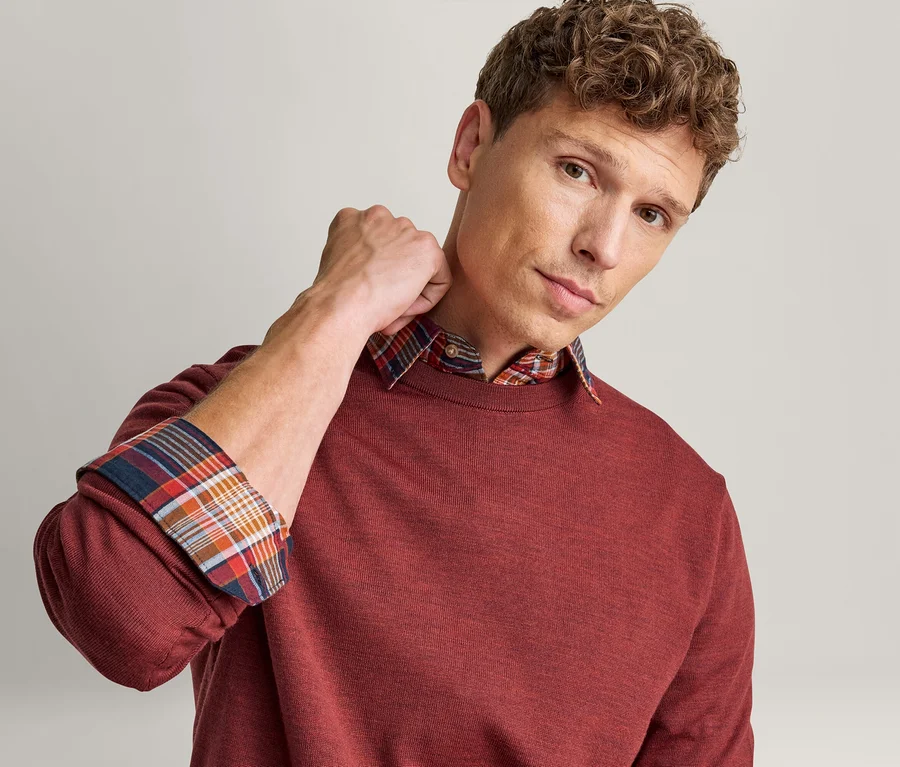 Mann trägt einen roten Pullover über einem karierten Hemd, wobei der Hemdkragen und die hochgekrempelten Ärmel sichtbar sind. Er blickt in die Kamera.