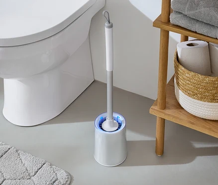 Im Badezimmer steht eine weiße Toilette neben einer WC-Bürste mit blauem Licht und einem Holzregal mit Handtüchern und Toilettenpapier.