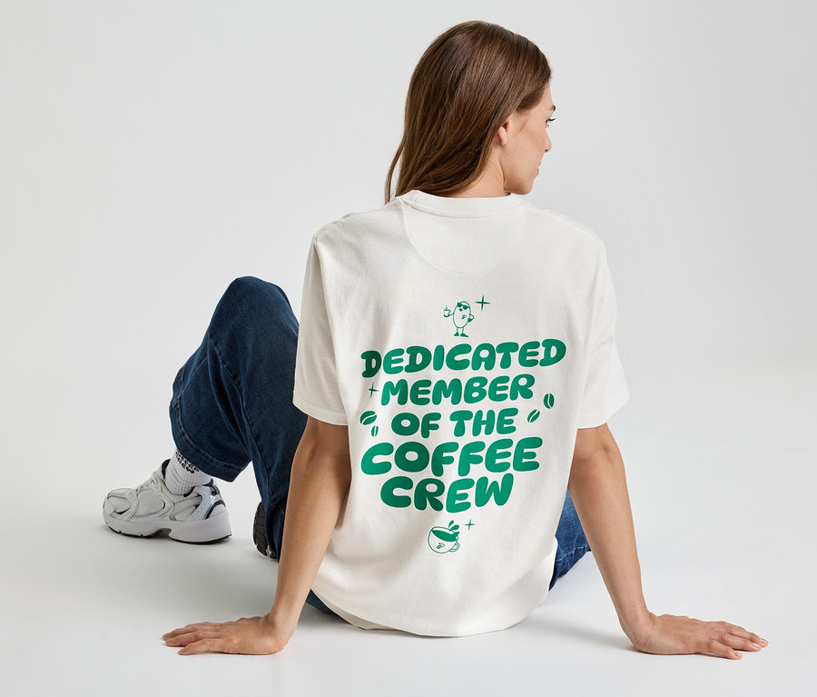 Frau sitzt auf dem Boden, trägt ein T-Shirt mit der Aufschrift "Dedicated Member of the Coffee Crew", dazu Jeans und Turnschuhe.