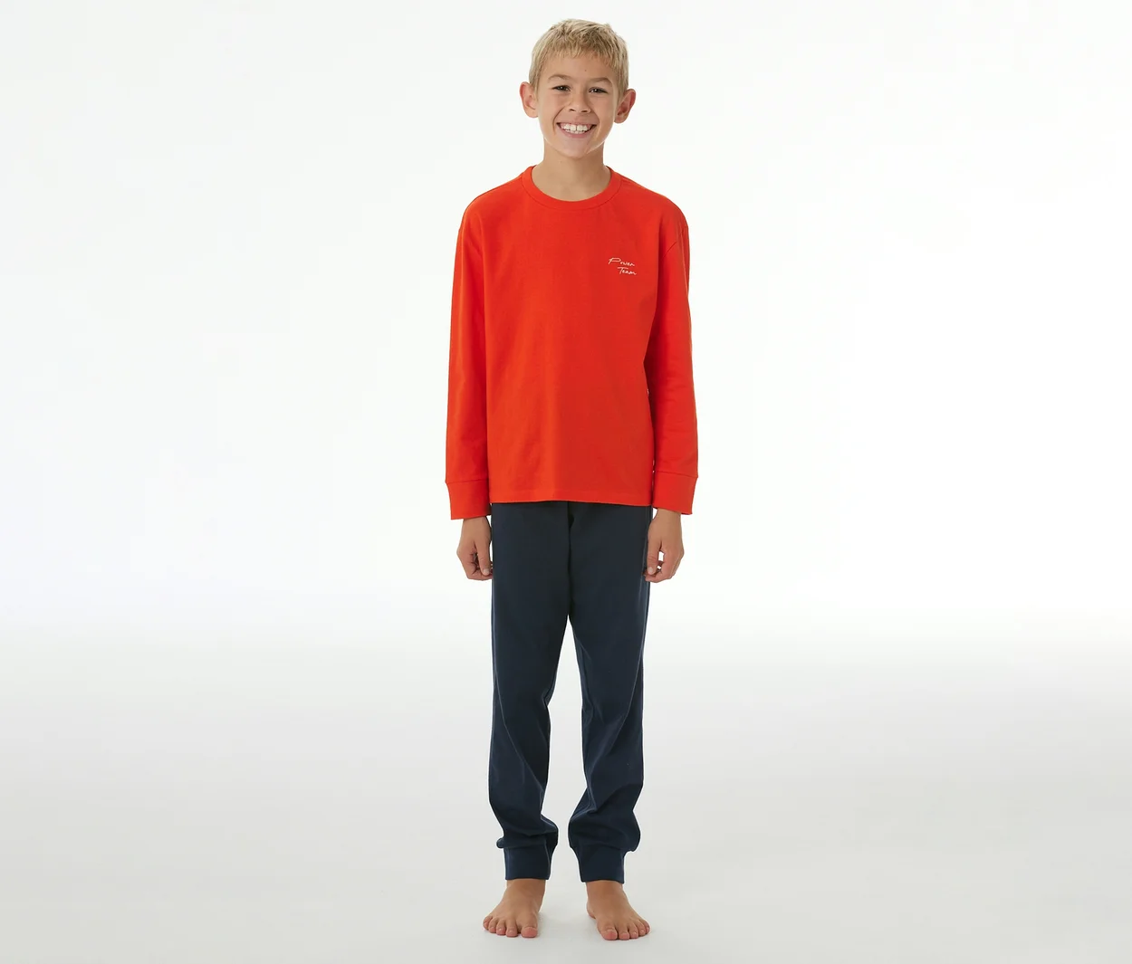 Lächelnder Junge in einem orangefarbenen SCHIESSER Kinder-Pyjama mit langen Ärmeln und dunkelblauer Hose mit Bündchen.