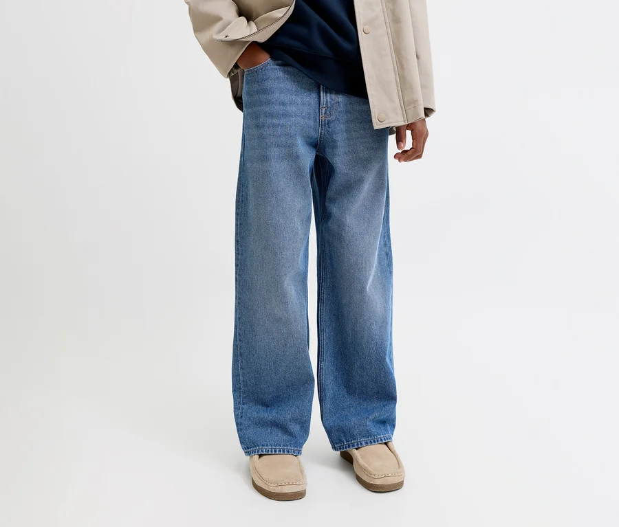 Person trägt weite blaue Jeans, eine beigefarbene Jacke, ein dunkelblaues Oberteil und beige Loafer. Eine Hand steckt in der Hosentasche.
