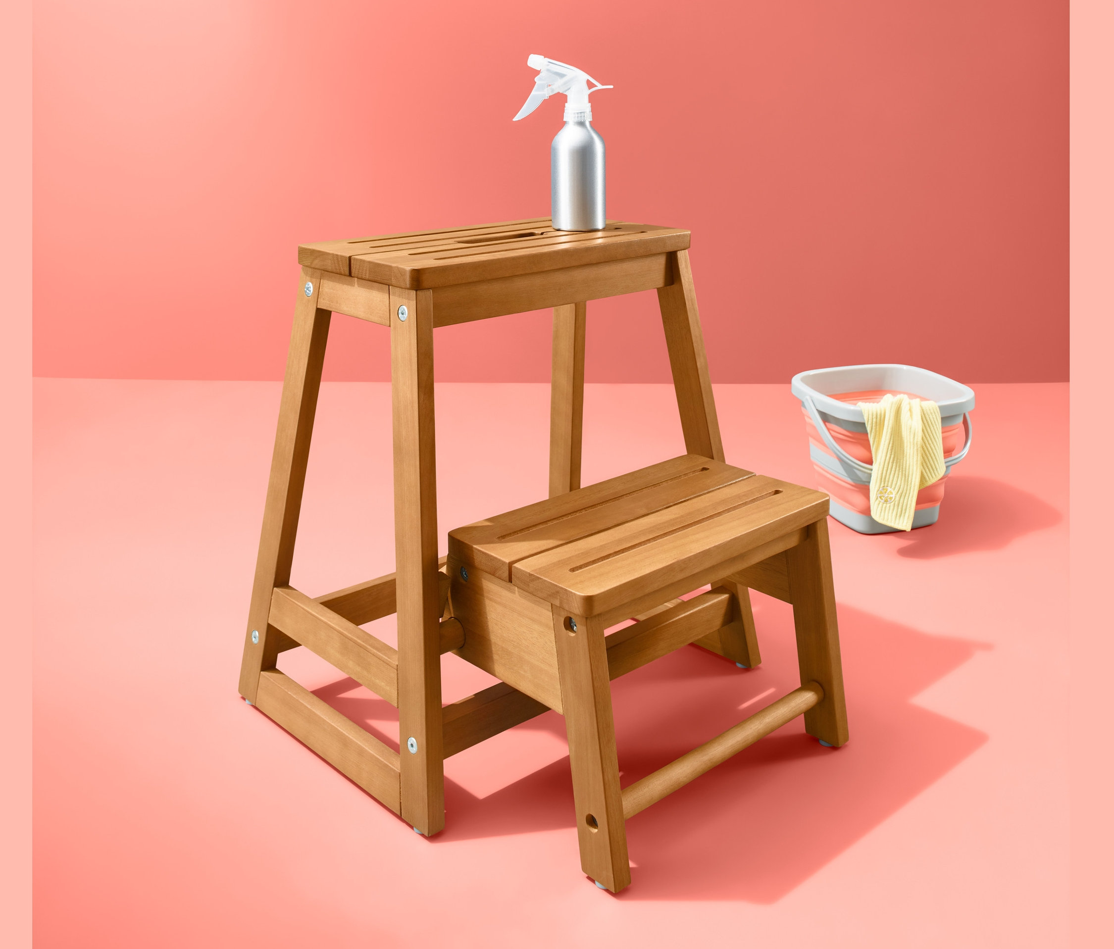 Ein hölzerner Klapptritt mit einer Sprühflasche und einem Eimer auf einem korallenroten Hintergrund.