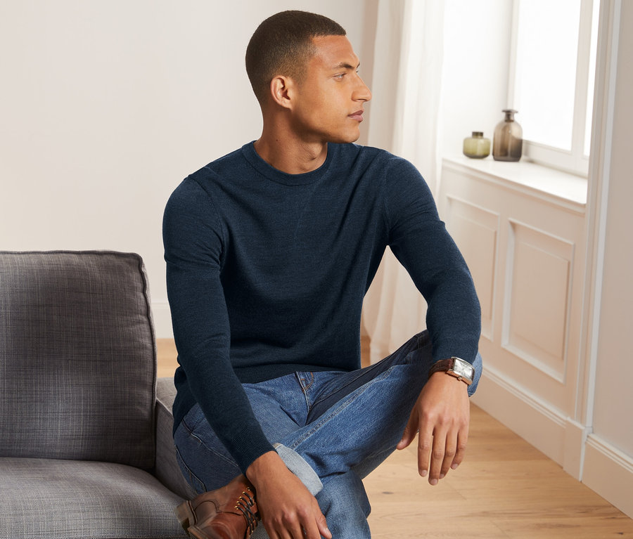 Mann sitzt auf grauem Sofa, trägt dunkelblauen Merino-Pullover mit Rundhalsausschnitt.