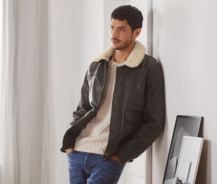 Mann an eine Wand gelehnt, trägt eine Lederjacke im Aviator-Style, einen Pullover und Jeans.