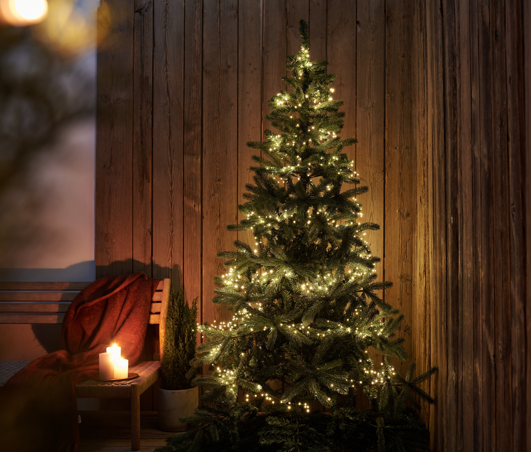 Ein beleuchteter Weihnachtsbaum mit XL-LED-Microlichterkette steht vor einer Holzwand. Daneben stehen auf einem Hocker brennende Kerzen und ein kleiner Baum im Topf.