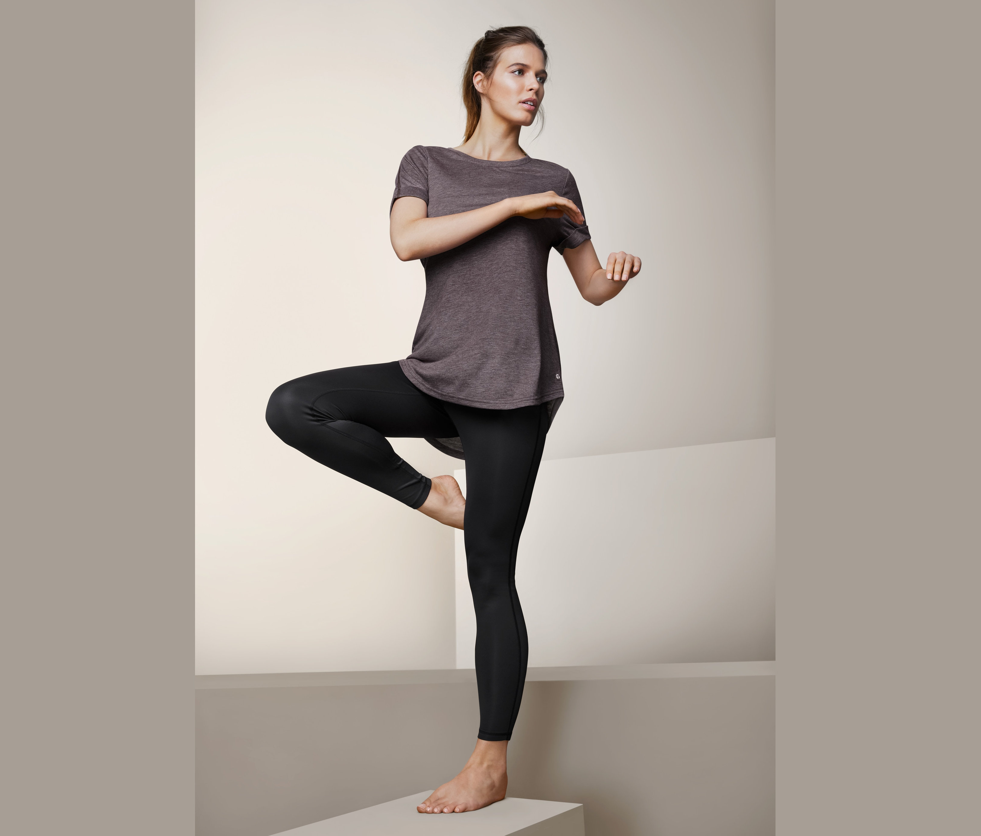 Frau steht auf einem Bein und trägt schwarze Leggings und ein Longshirt.